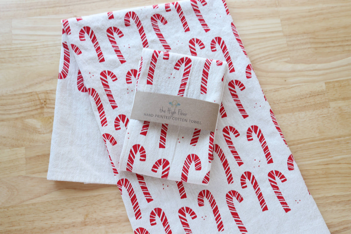 A folded white cotton towel with a red candy cane pattern and a kraft paper label, placed on a light wood surface.