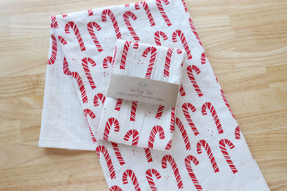 A folded white cotton towel with a red candy cane pattern and a kraft paper label, placed on a light wood surface.