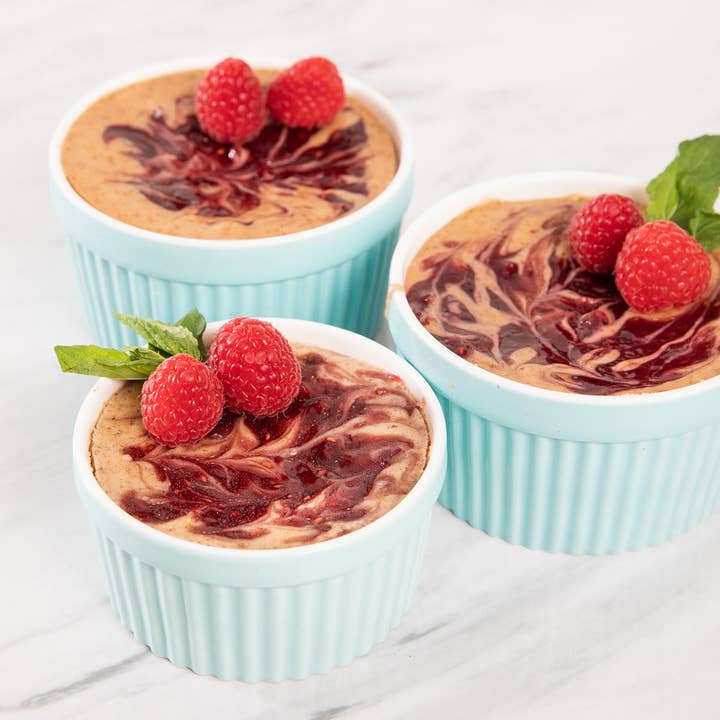 Three light blue ramekins filled with a chocolate mousse and raspberry preserves garnished with raspberries on a light background.