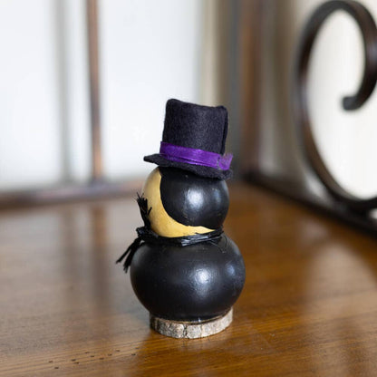 A small decorative figurine with a black body, yellow face, black hat, and purple band, standing on a wooden surface in an indoor setting.