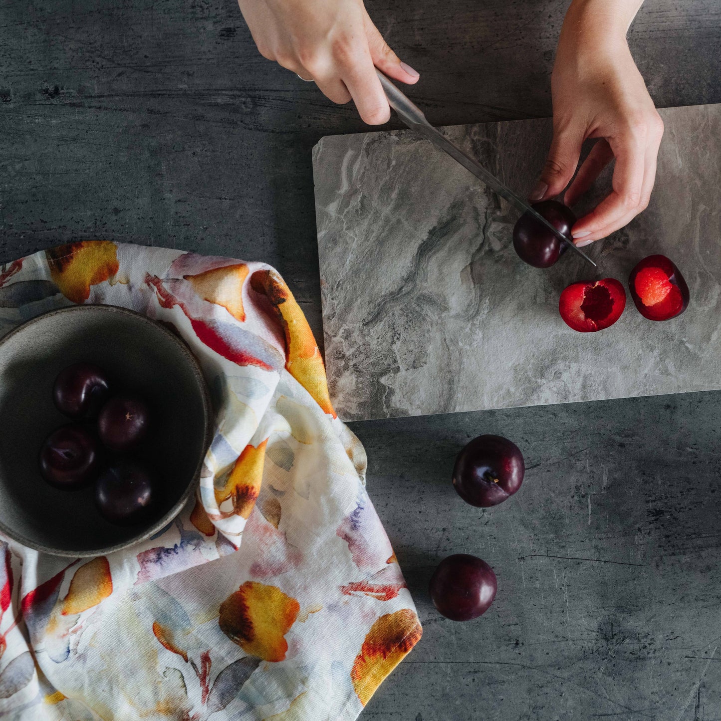 Person cutting plums on a marble slab with a bowl of plums and floral tea towel nearby.