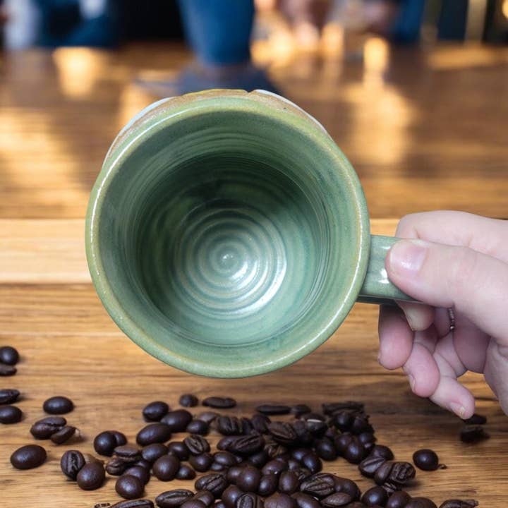Green ceramic mug held over a wooden surface with coffee beans