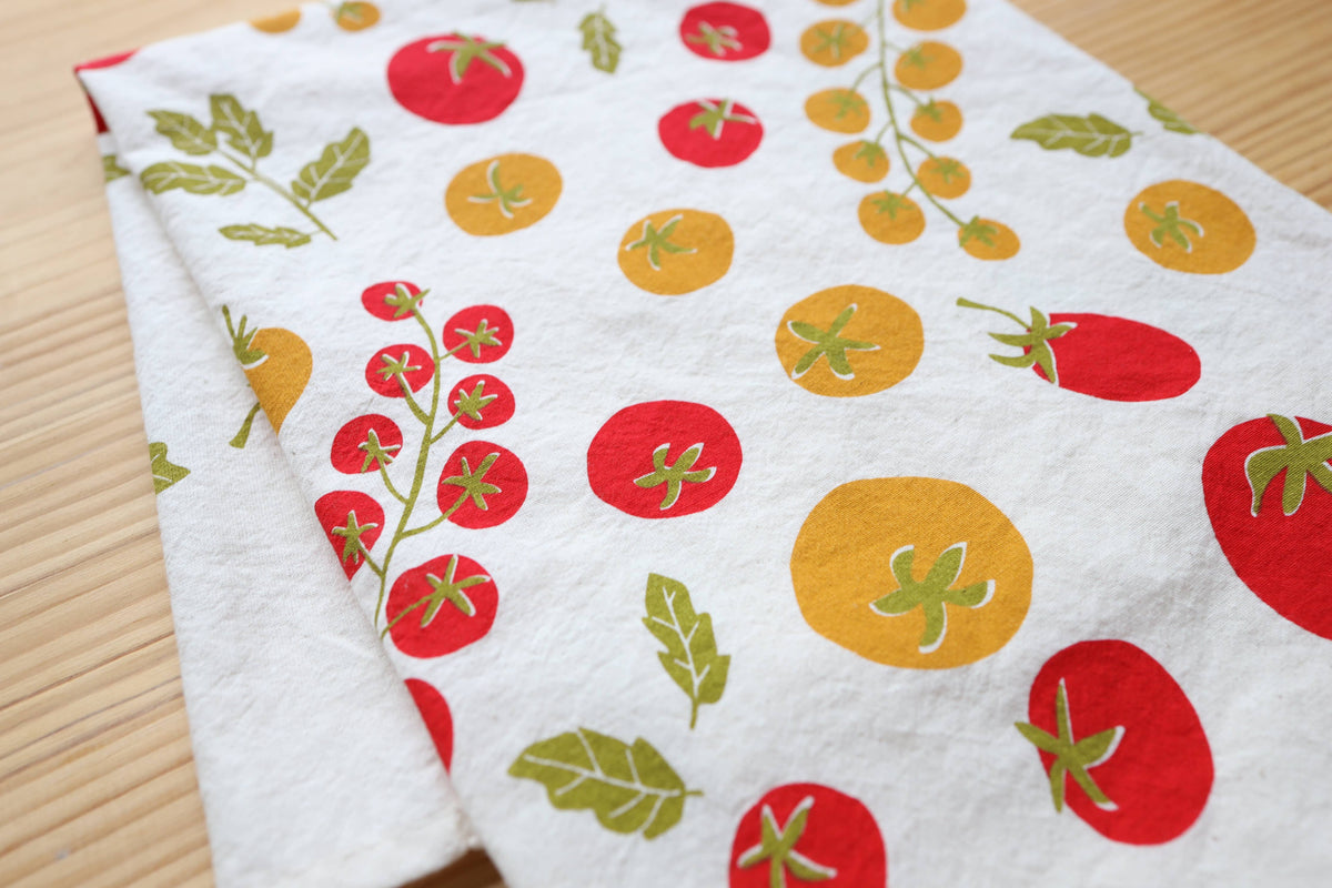 A white kitchen towel featuring a colorful print of tomatoes and green leaves, laid flat on a light wood surface.