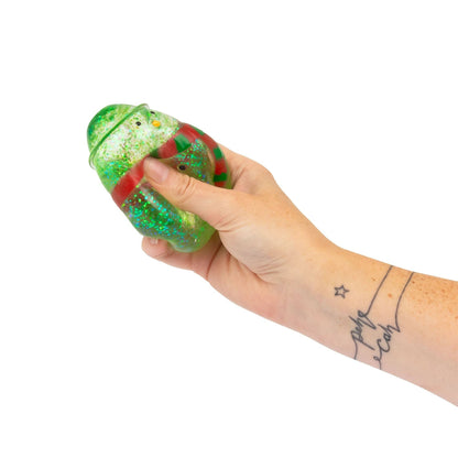 A hand holding a green glittery snowman shaped squishy toy with red accents, shown against a plain white background.