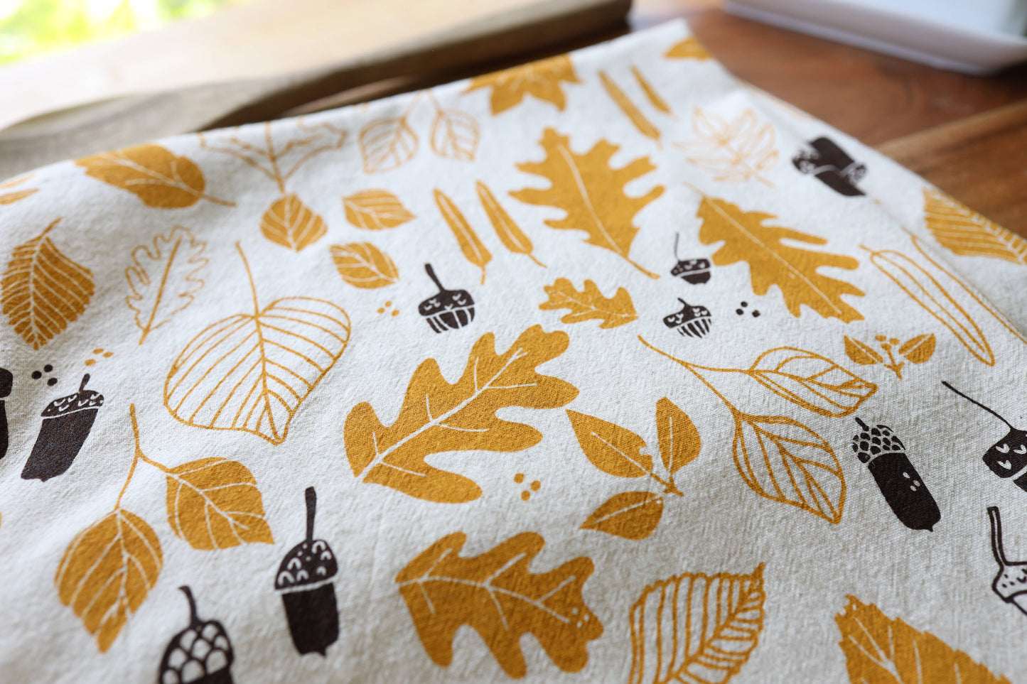 A close-up of a white tea towel, printed with a pattern of autumn leaves and acorns in mustard yellow and dark brown.