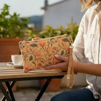 A person holding a rectangular clutch bag with a vintage floral pattern in warm orange, red, and green tones on a beige background. The clutch has a cork wrist strap and gold-tone hardware, set in an outdoor patio environment with a wooden table, coffee cup, and potted plants in the background.