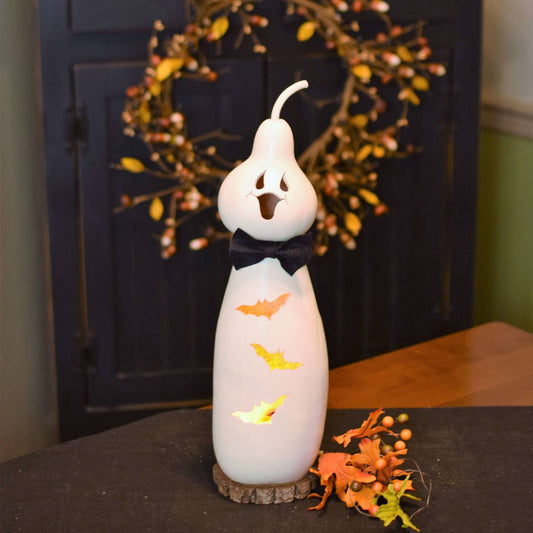 A white ceramic Halloween ghost lantern gourd, decorated with a black bow tie, orange bat cutouts, and a glowing light inside, displayed on a dark tabletop with autumn leaves and a wreath in the background.
