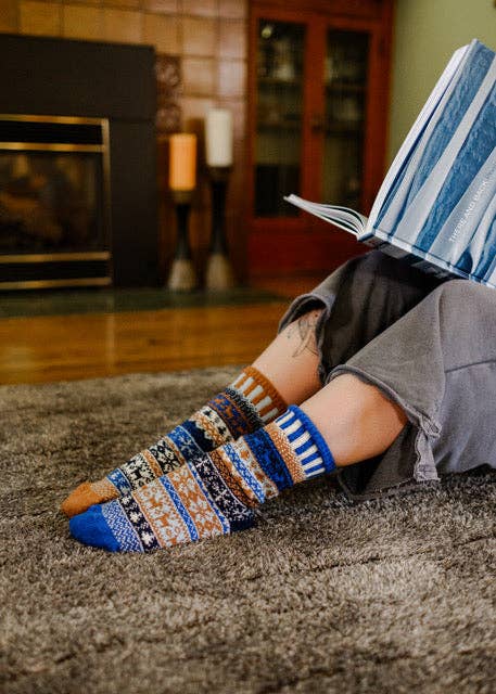 A person sitting on a shag rug in a cozy living room, wearing a pair of multicolored patterned crew socks while holding an open book.