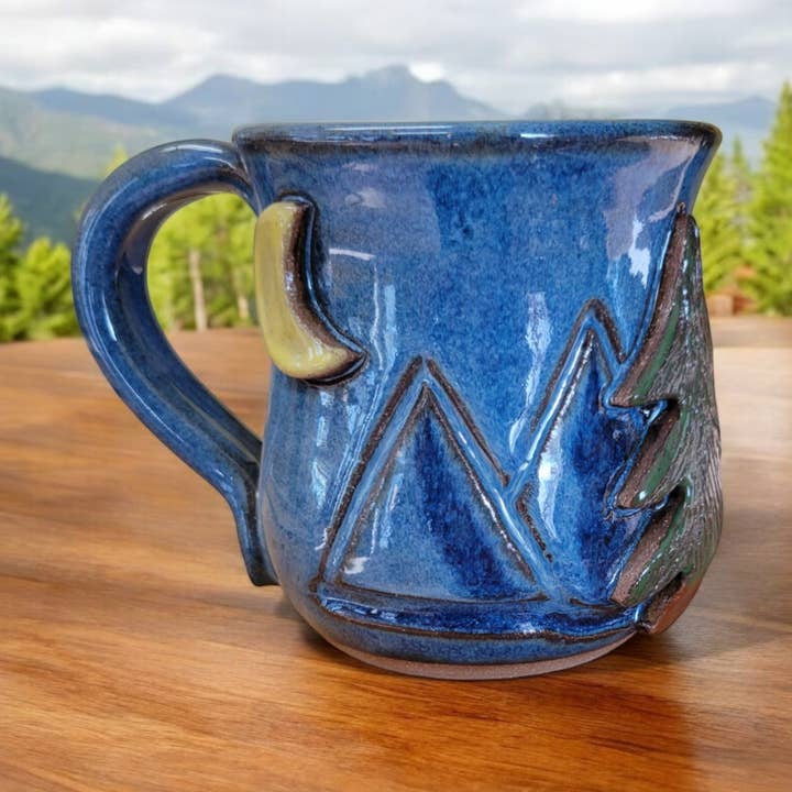 Blue ceramic mug with mountain design on a wooden surface, mountainous landscape in the background