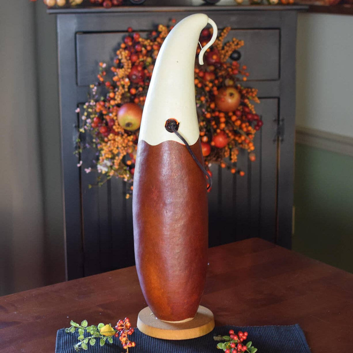 A decorative gourd sculpture featuring a smooth reddish-brown body and a curved cream-colored head, displayed on a dark wood table with a holiday wreath in the background.