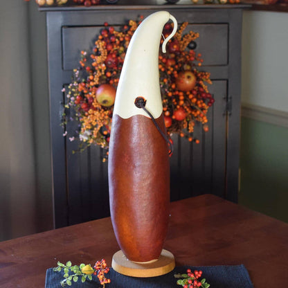 A decorative gourd sculpture featuring a smooth reddish-brown body and a curved cream-colored head, displayed on a dark wood table with a holiday wreath in the background.