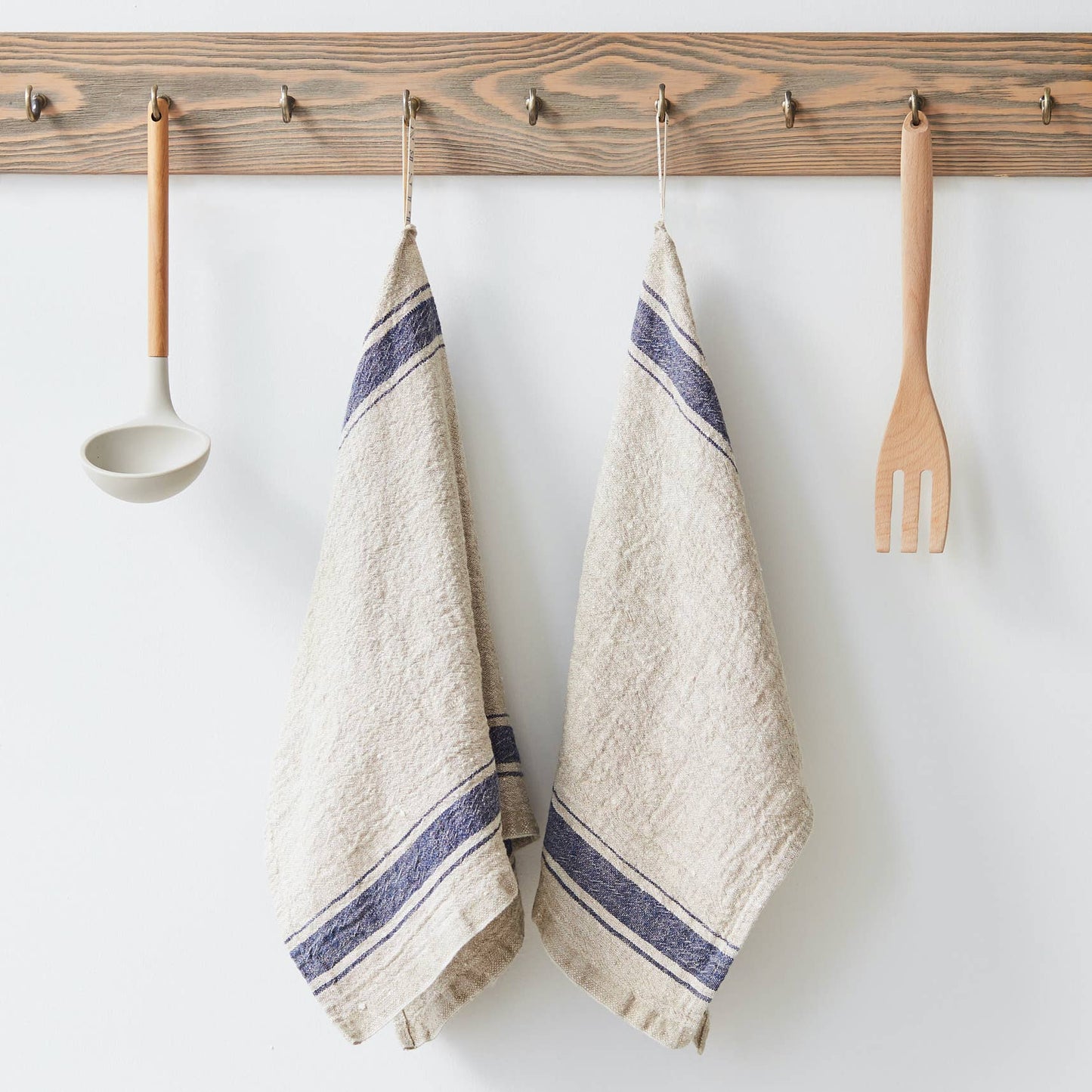 Two beige towels with blue stripes hanging on a wooden rack with kitchen utensils.