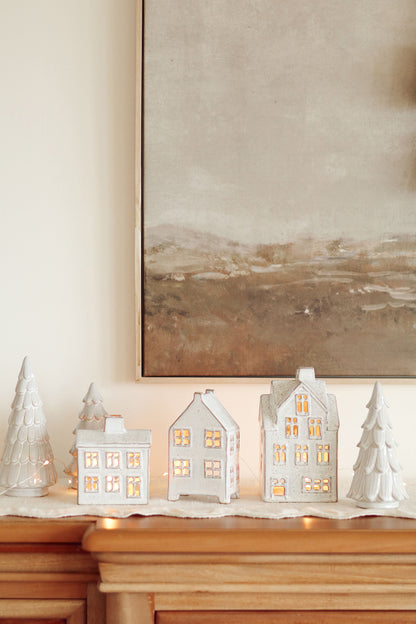 Decorative ceramic houses and trees with warm lighting on a mantelpiece, with a painting in the background.