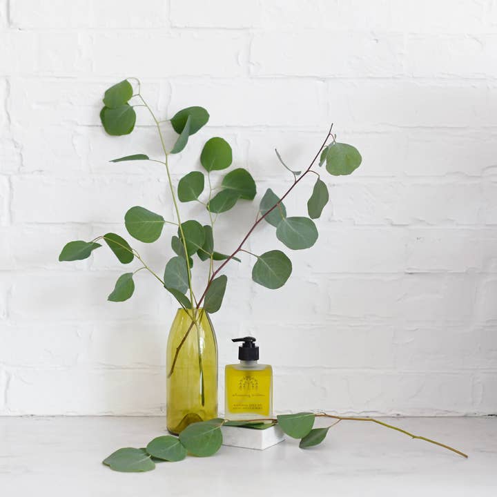 Bottle of body oil with green leaves on a white surface against a white brick wall.