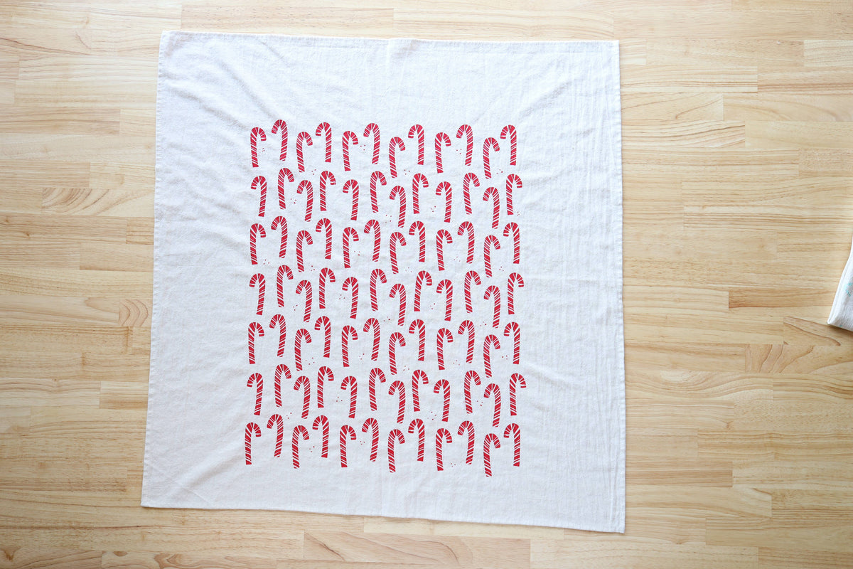 A white tea towel featuring a grid pattern of red and white candy canes, laid flat on a light wood table.