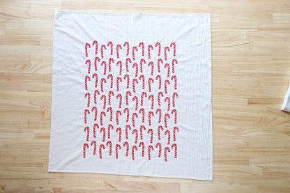 A white tea towel featuring a grid pattern of red and white candy canes, laid flat on a light wood table.