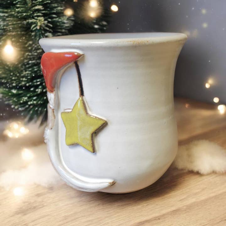 White ceramic mug with red Santa hat and yellow star on a wooden surface with blurred Christmas tree and lights in the background.
