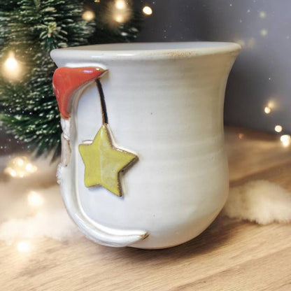 White ceramic mug with red Santa hat and yellow star on a wooden surface with blurred Christmas tree and lights in the background.
