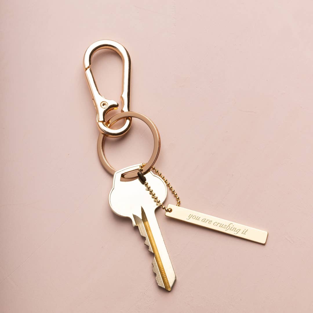 Gold keychain with a key-shaped charm and motivational tag on a pink background