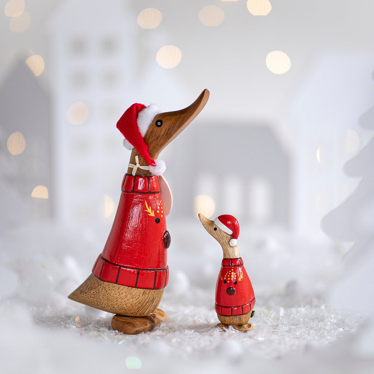 Wooden duck figurines wearing red outfits and hats on a snowy background with blurred lights.