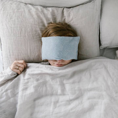 Person lying in bed with an eye pillow on on, partially covered by a blanket.