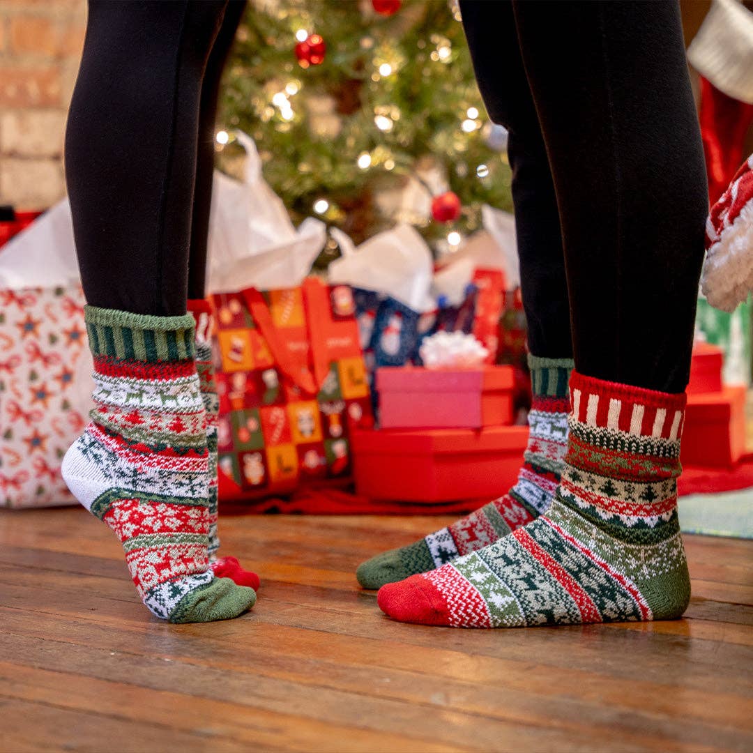 A person wearing black leggings and festive Christmas knit socks with red, green, white, and blue patterns, standing on a wooden floor in front of a decorated Christmas tree and wrapped gifts.