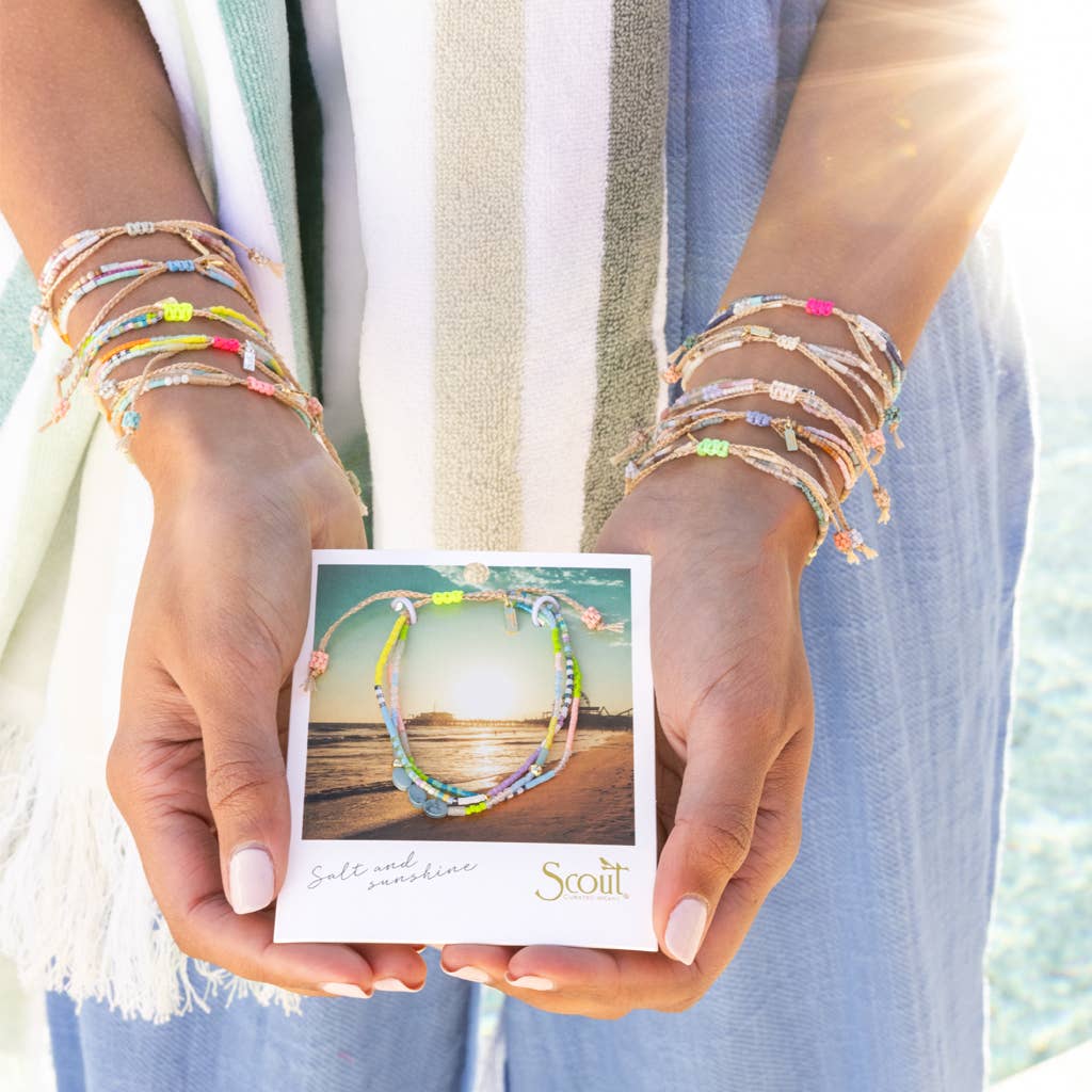 Person wearing colorful bracelets holding a card with a bracelet design, against a blurred natural background.