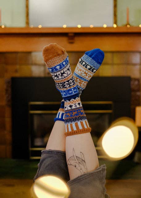 A person's legs are raised in the air, showing a pair of patterned crew socks with blue, burnt orange, and white Fair Isle designs, set against a cozy fireplace background.