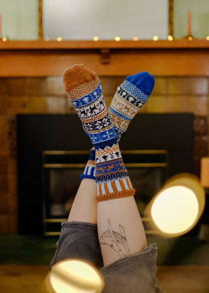 A person's legs are raised in the air, showing a pair of patterned crew socks with blue, burnt orange, and white Fair Isle designs, set against a cozy fireplace background.