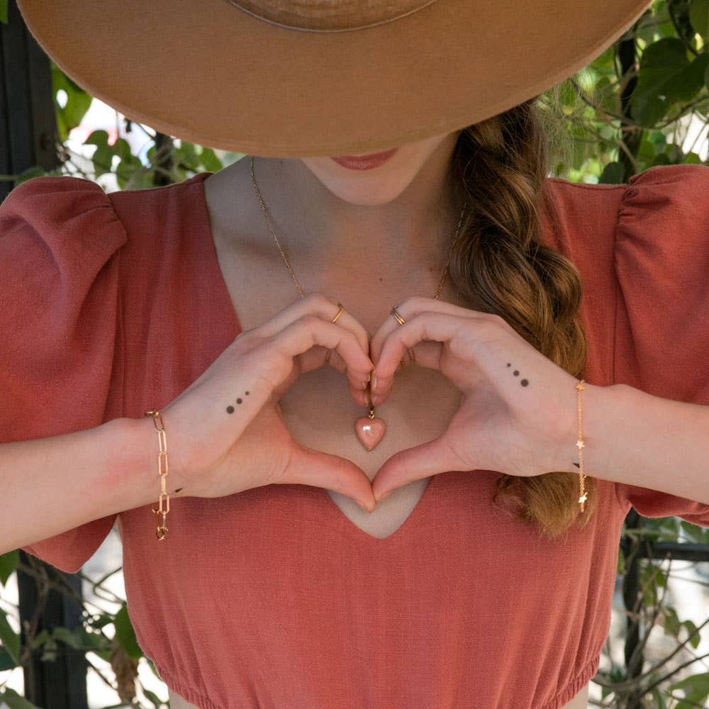 Person wearing a wide-brimmed hat and pink top, making a heart shape with their hands.