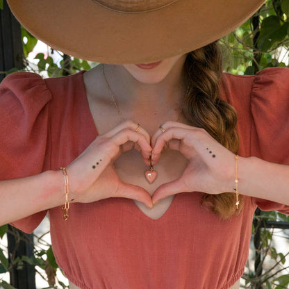 Person wearing a wide-brimmed hat and pink top, making a heart shape with their hands.