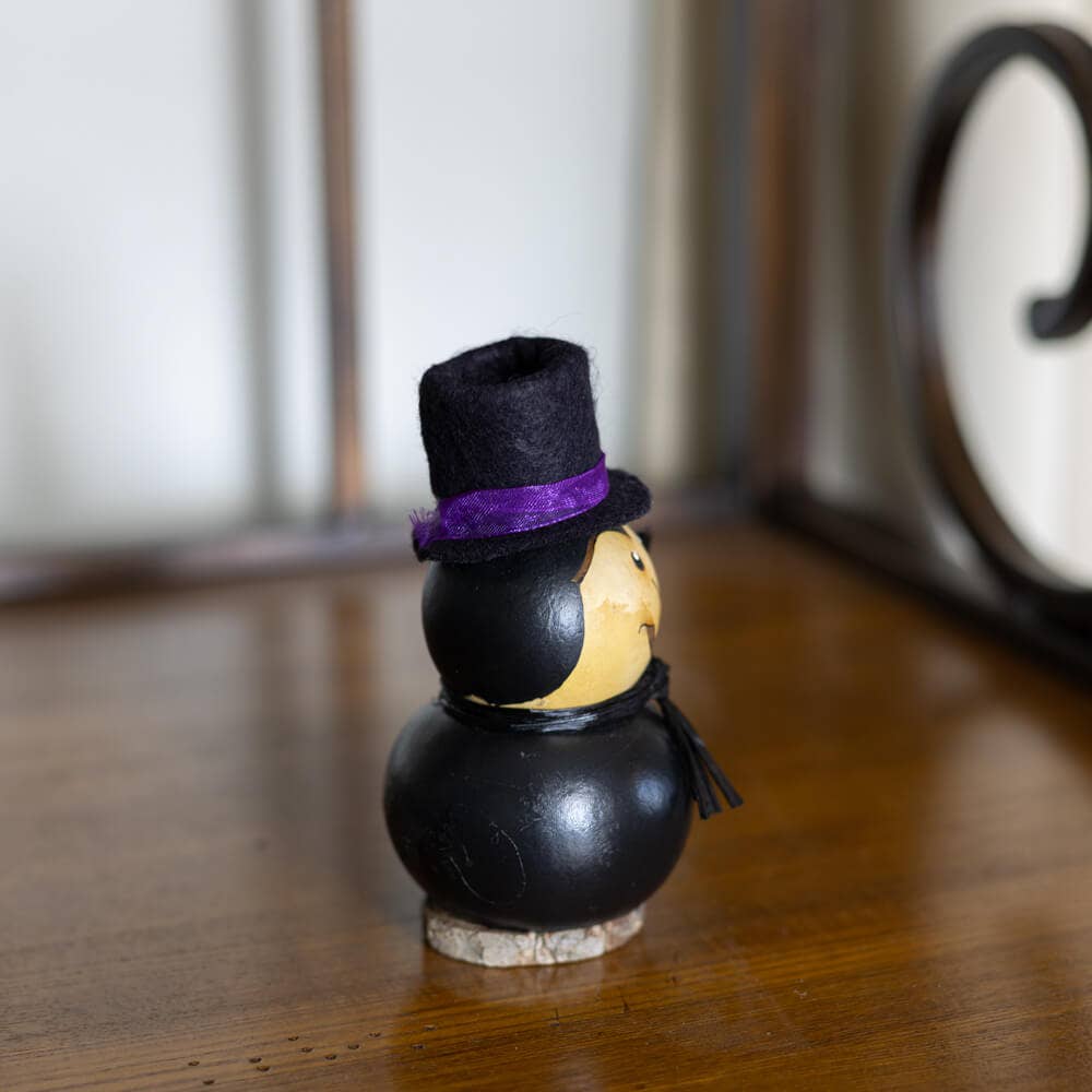 A small decorative figurine with a black body, yellow face, black hat, and purple band, standing on a wooden surface.