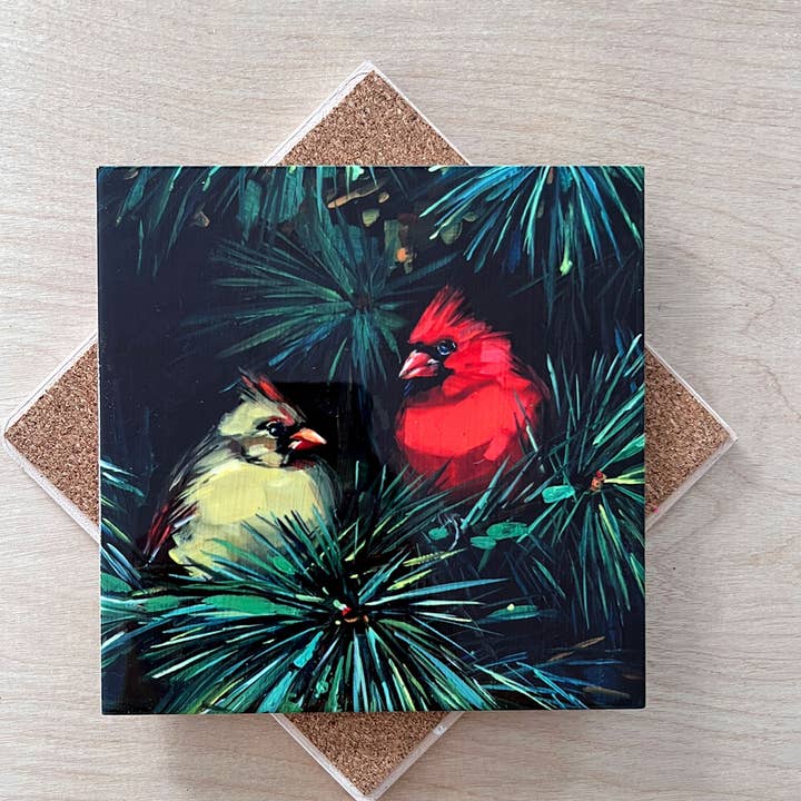 Trivet with painting of two cardinals in a pine tree on a wooden surface.