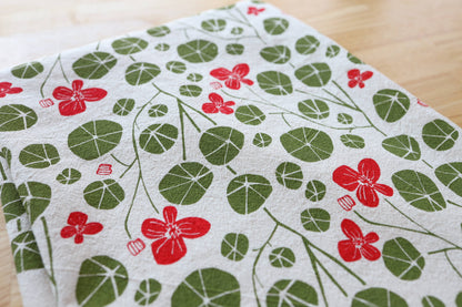 A close-up of a white fabric kitchen towel featuring a repeating pattern of green round leaves and red flowers, laid flat on a light wood surface.