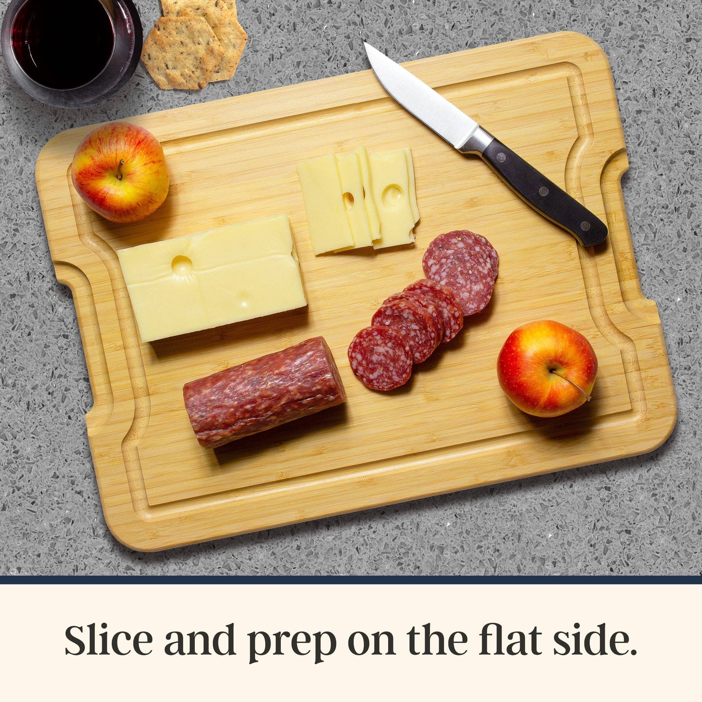 Wooden cutting board with sliced cheese, salami, and apples on a gray surface. Text at bottom: Slice and prep on the flat side.