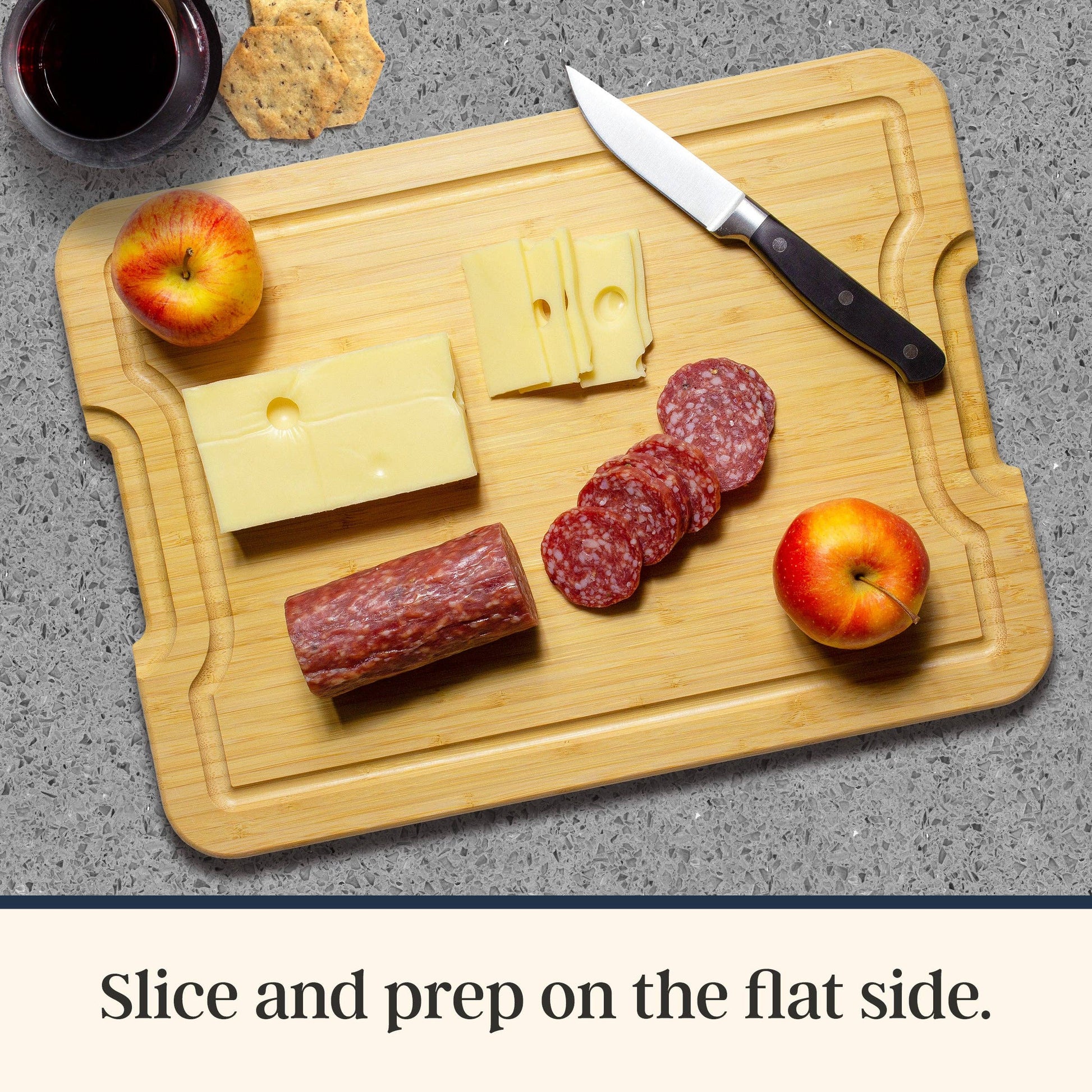 Wooden cutting board with sliced cheese, salami, and apples on a gray surface. Text at bottom: Slice and prep on the flat side.