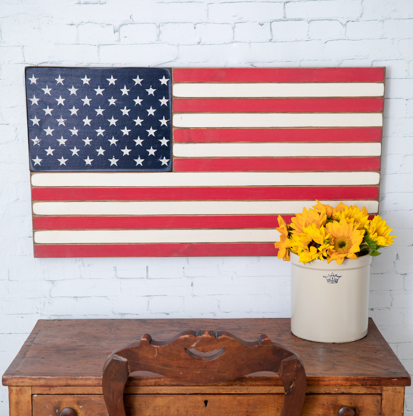 Wooden American flag on a white brick wall with a wooden chest and yellow flowers in the foreground.