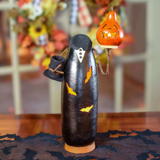 A Halloween decoration shaped like a headless jack o lantern with a black hat, and a small orange gourd lantern on a stand, displayed on a table with a blurred autumn background.