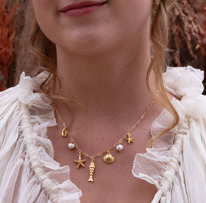 Close-up of a person wearing a gold necklace with starfish, shell, and pearl charms against a blurred natural background.