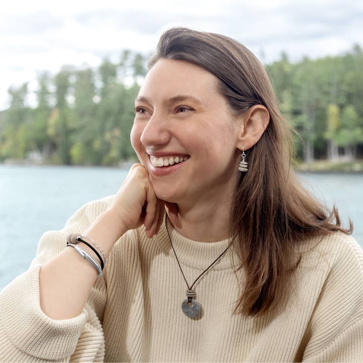 Woman smiling outdoors by a lake with trees in the background. She is wearing stone earrings, a necklace, and bracelets.