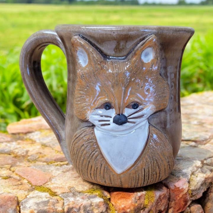 Ceramic mug with fox face design on a stone surface with a green field background