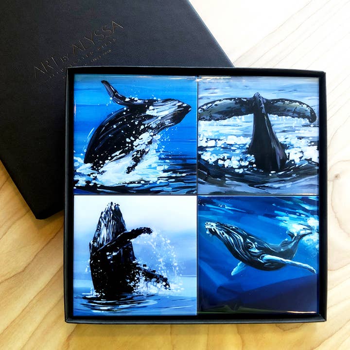 four ceramic coasters of whales in water on a wooden surface in a gift box.