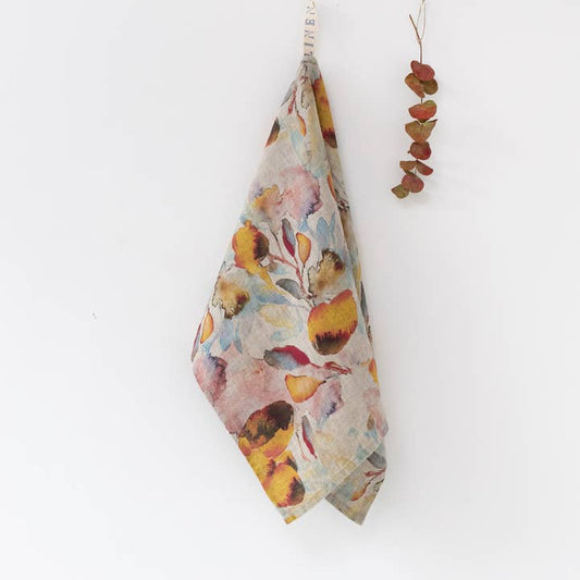 A hanging fabric tea towel with a watercolor-style floral print in soft pink, yellow, orange, and blue tones on a white background, with a small dried leaf sprig hanging to the right.
