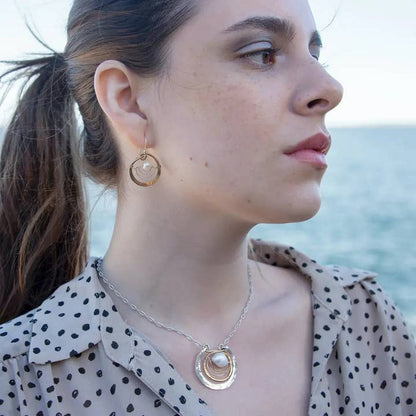 Woman wearing gold hoop earrings and a necklace with a pendant, standing by the ocean.