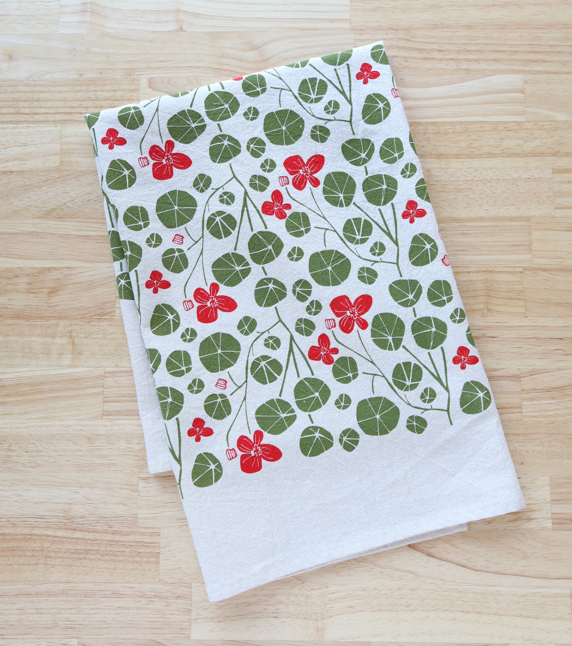 A folded white kitchen towel with a green and red nasturtium leaf and flower pattern, displayed on a light wood grain surface.
