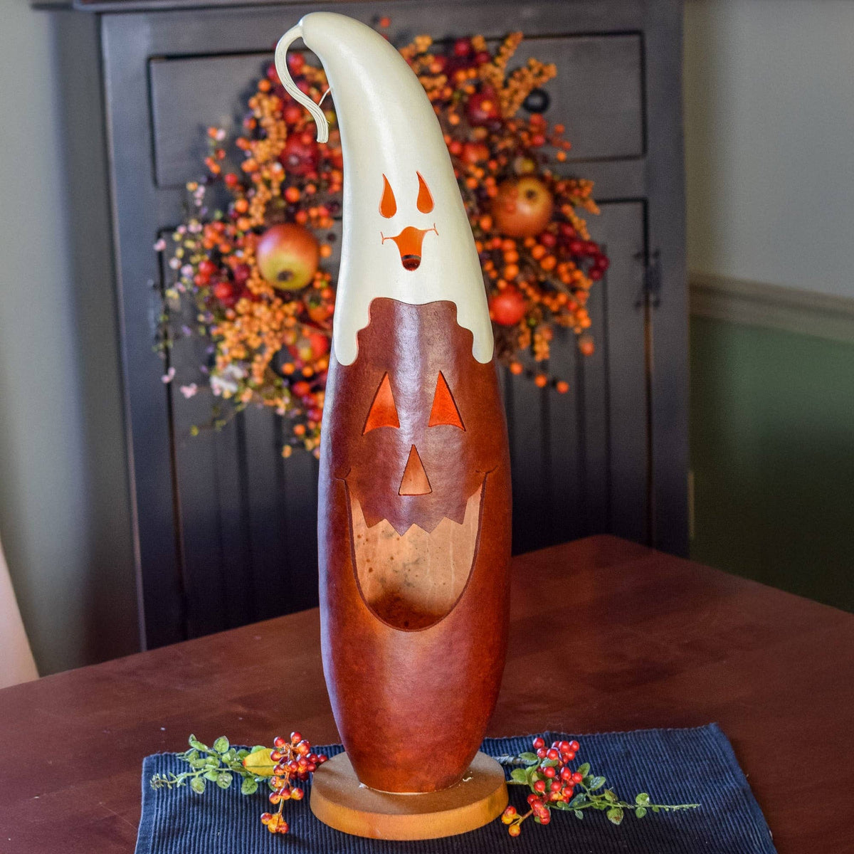 A carved wooden jack-o'-lantern luminary with a white ghost-shaped top and orange cutout details, displayed on a dark table runner with a fall wreath in the background.