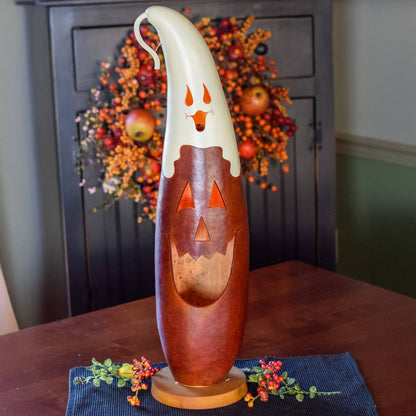 A carved wooden jack-o'-lantern luminary with a white ghost-shaped top and orange cutout details, displayed on a dark table runner with a fall wreath in the background.