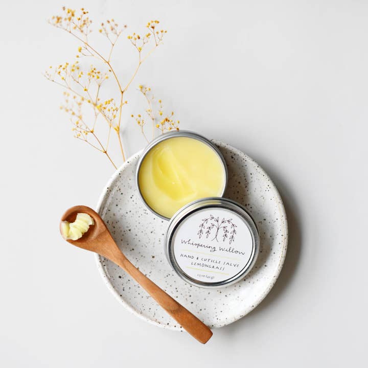 Open tin of salve on a speckled ceramic plate with a wooden spoon and dried flowers.
