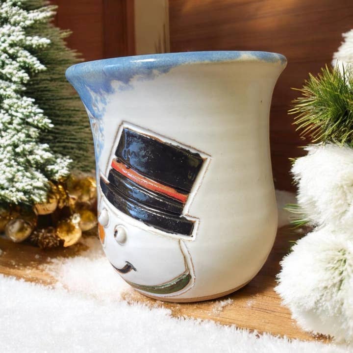 Snowman-themed ceramic mug with a black top hat on a snowy surface with Christmas decorations.