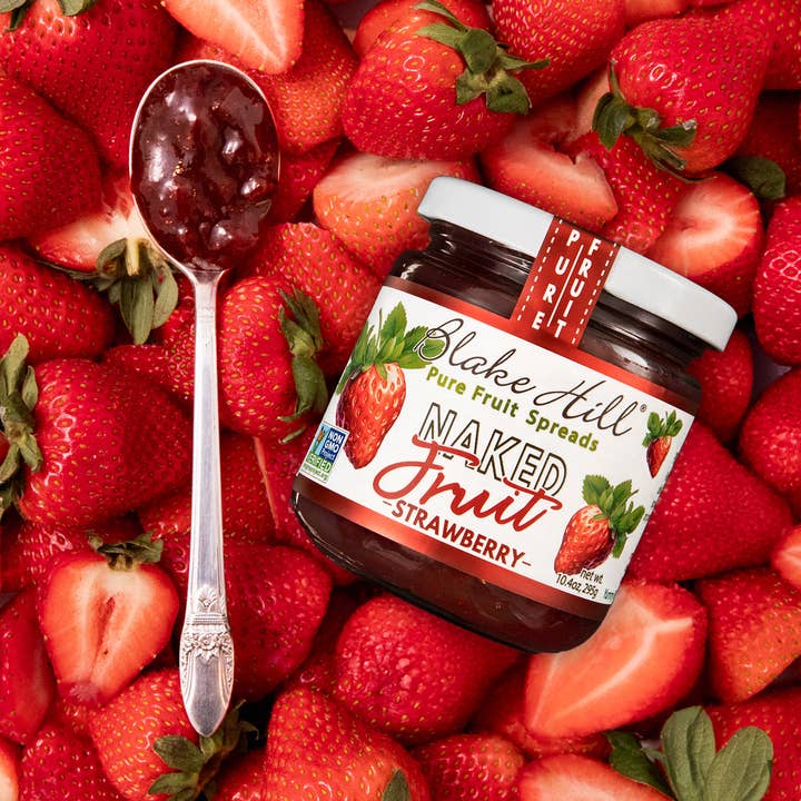 A jar of Blake Hill Strawberry preserves on a bed of strawberries with a spoonful of preserves.